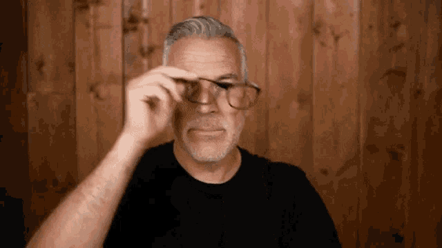 a man in a black shirt is adjusting his glasses against a wooden wall .