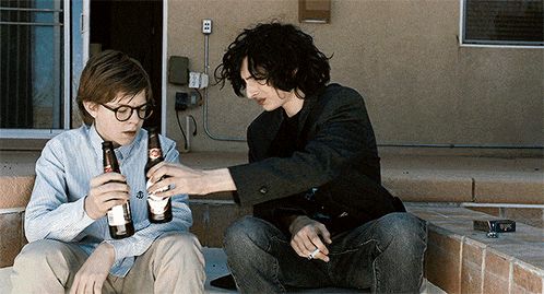 two boys sit on a porch drinking beer and smoking a cigarette