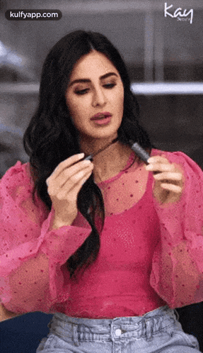 a woman in a pink top and jeans is holding a lipstick in her hand .
