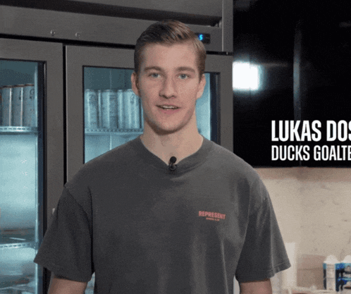 a man wearing a lukas do ducks goalie t-shirt stands in front of a refrigerator
