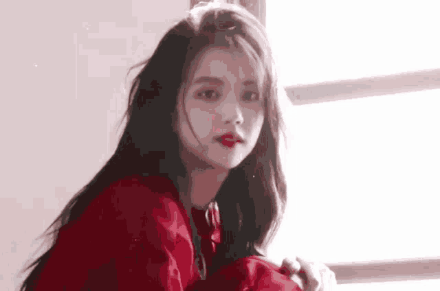 a girl in a red dress is sitting in front of a window and looking at the camera .