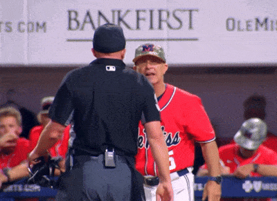 a baseball player in a red jersey talks to an umpire in front of a bankfirst sign