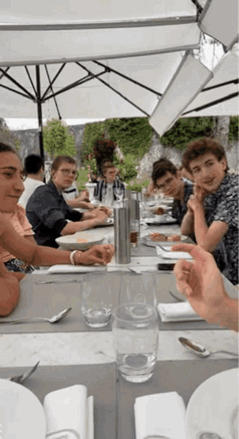 a group of people are sitting at a table under an umbrella eating food