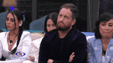 a man with a beard sits in front of a group of women with the words grandefratello on the bottom