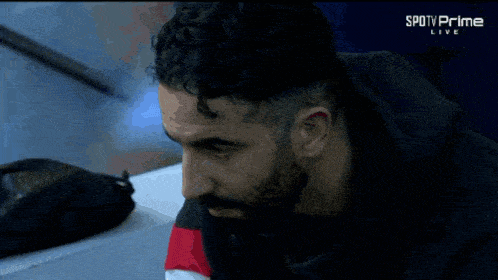 a man with a beard sits in front of a screen that says spotv prime