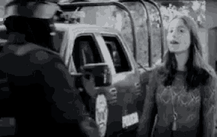 a black and white photo of a woman talking to a police officer in front of a police truck .
