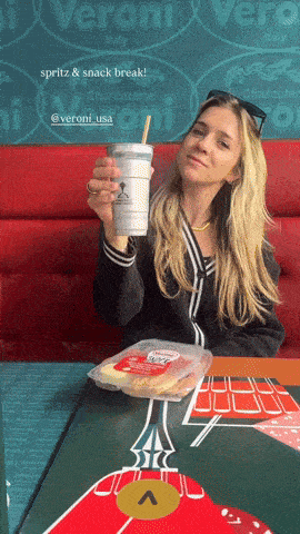a woman sitting at a table holding a drink in front of a sign for veroni