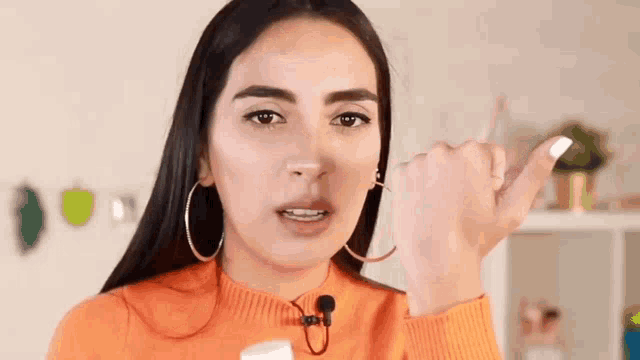 a woman wearing hoop earrings and an orange sweater is talking into a microphone and making a funny face .