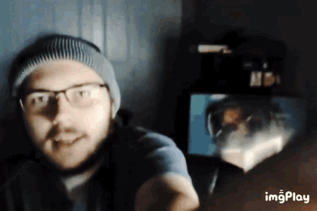 a man wearing glasses and a beanie is standing in front of a computer screen with a reflection of himself