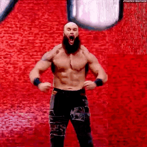 a wrestler without a shirt is standing in front of a red wall with his mouth open .