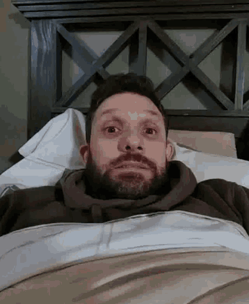 a man with a beard is laying in a bed with his eyes closed and a surprised look on his face .