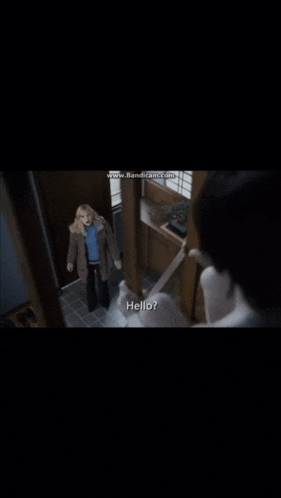 a woman in a blue sweater is standing in a hallway talking to a man in a white shirt who says hello