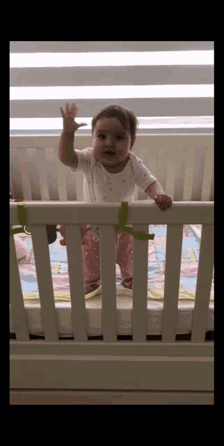 a baby is standing in a white crib and waving at the camera