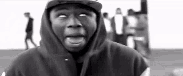 a black and white photo of a man wearing a hooded jacket and a hat making a funny face .