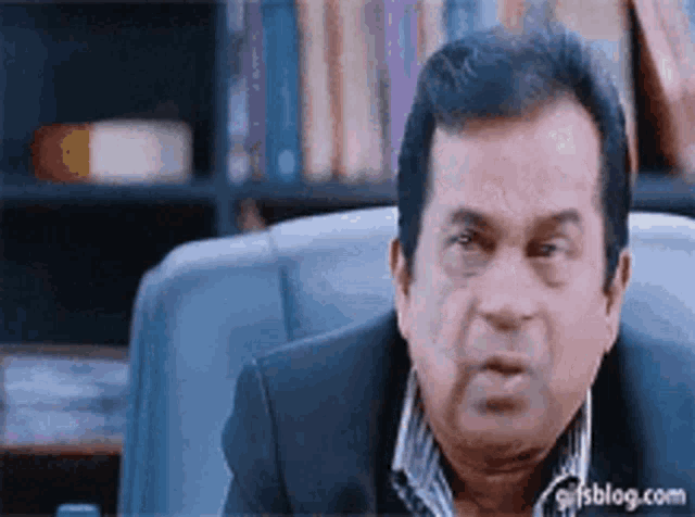 a man in a suit is sitting in a chair in front of a bookshelf and making a funny face .