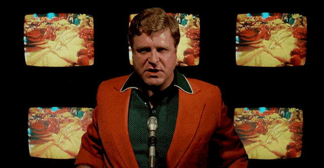 a man in an orange jacket is speaking into a microphone in front of a row of televisions