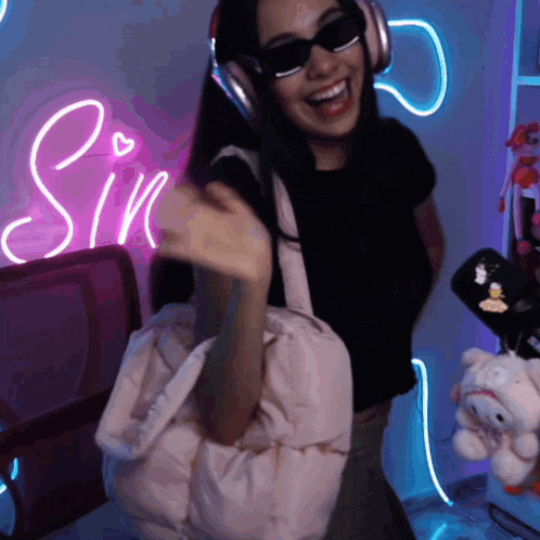 a woman wearing headphones and sunglasses holds a pink bag in front of a sin sign