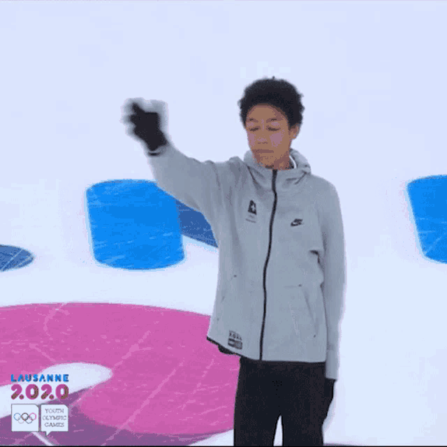 a young man in a grey nike jacket stands in front of a lausanne 2020 sign