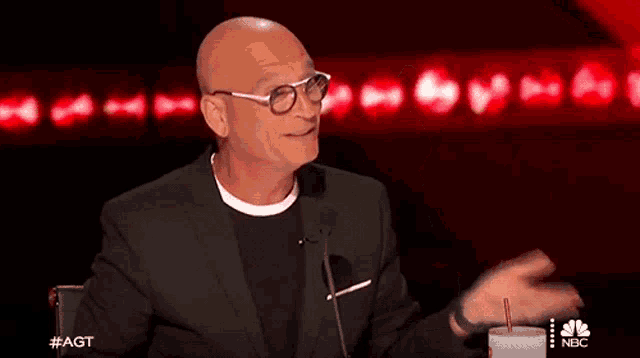a bald man wearing glasses and a black suit is sitting in front of a microphone .