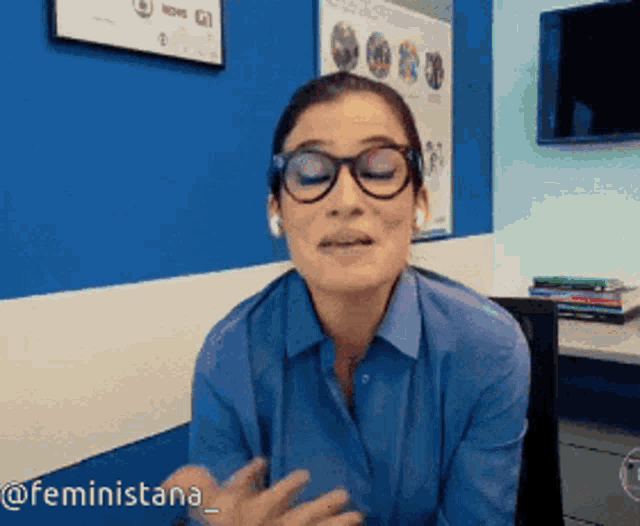 a woman wearing glasses and a blue shirt is sitting in front of a blue wall with the hashtag feministana on the bottom