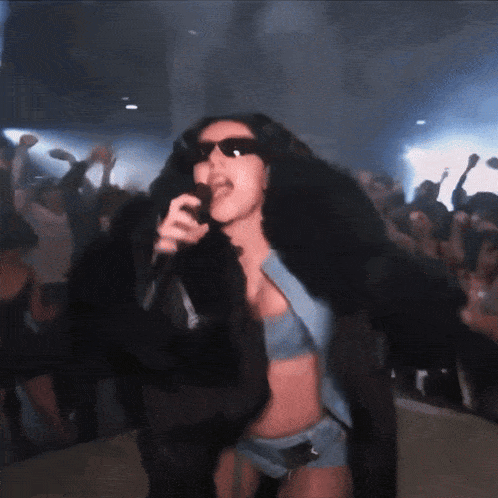 a woman wearing sunglasses and a fur coat sings into a microphone