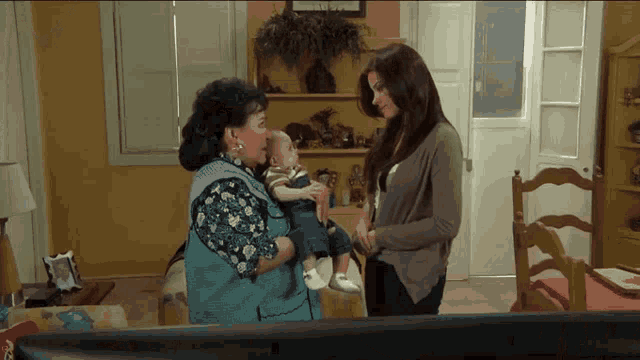 a woman holds a baby while another woman looks on in a living room