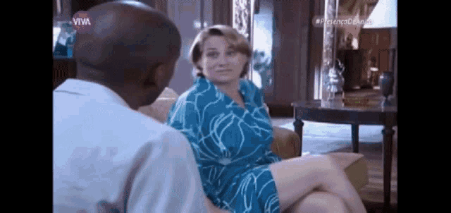 a woman in a blue dress sits on a couch talking to a man in a living room .