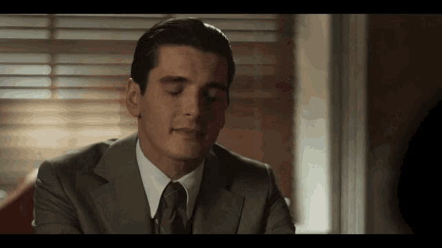 a man in a suit and tie is sitting in front of a window with his eyes closed and his mouth open .