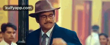 a man wearing a hat and glasses is standing in a room with other men .