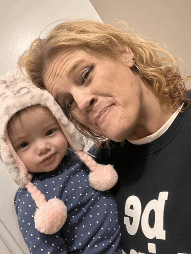 a woman is holding a baby wearing a hat and a black shirt that says pe