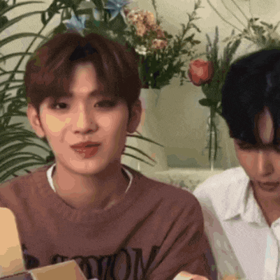 two young men are sitting next to each other in front of flowers . one of the men is holding a box .