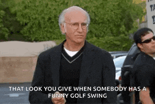 a man in a suit and sweater is standing in a parking lot with a funny golf swing .
