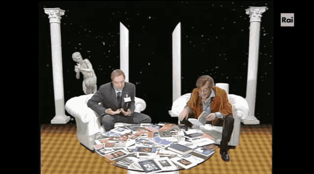two men sit in chairs in front of a pile of magazines and a sign that says rai on it