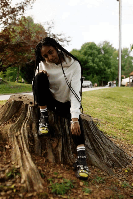 a woman is sitting on a tree stump wearing a white hoodie and black pants