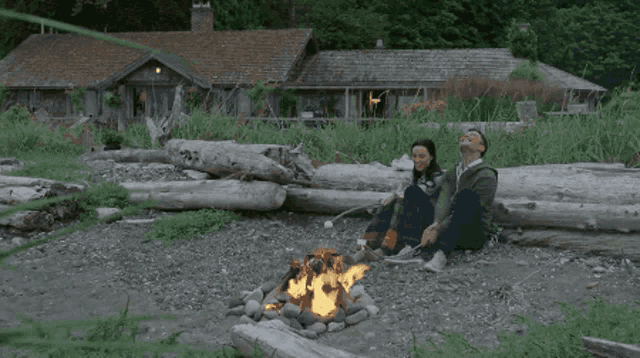 a man and a woman are sitting around a campfire in front of a house