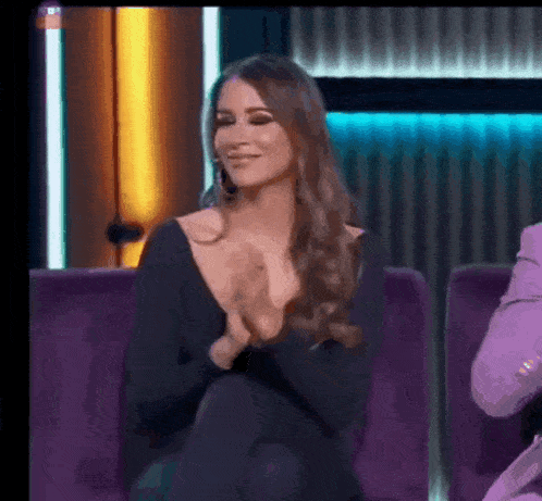 a woman in a black dress is sitting on a purple couch and clapping her hands .