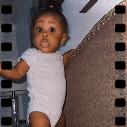 a baby in a white bodysuit is standing in front of a chair