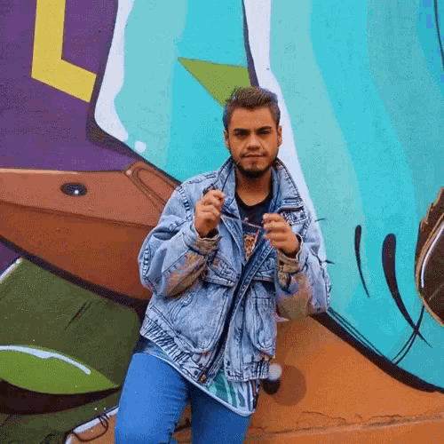 a man in a denim jacket is standing in front of a wall with graffiti on it