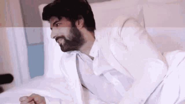 a man with a beard is laying in bed wearing a white shirt and tie .