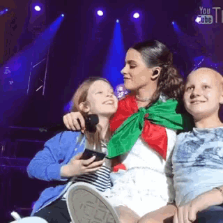 a woman is sitting on a stage with two children and holding a cell phone .