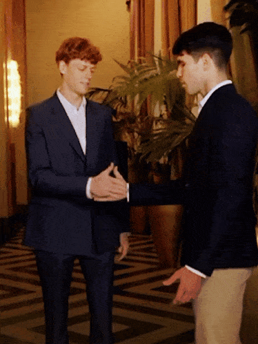 two men in suits shake hands in front of a plant