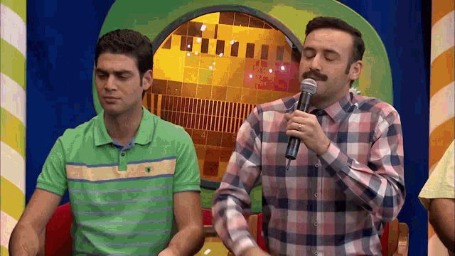 a man in a plaid shirt holds a microphone while another man looks on