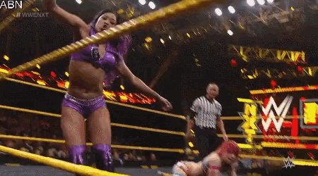 a woman in purple shorts is standing in a wrestling ring while a referee looks on .