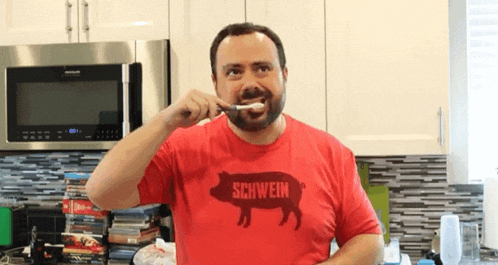 a man is brushing his teeth in a kitchen wearing a schwein shirt