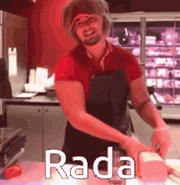 a man in a red shirt and black apron is standing in a kitchen cutting meat .
