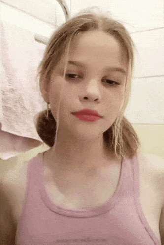 a young girl in a pink tank top looks at the camera with a towel in the background