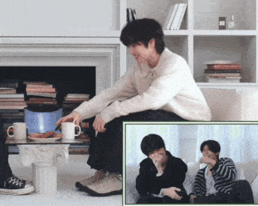 a man in a white sweater sits in front of a fireplace holding a white mug