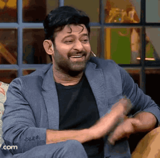 a man in a suit and black shirt is sitting on a couch and laughing