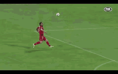 a soccer player in a red uniform is kicking the ball while another player in a green uniform lays on the ground .