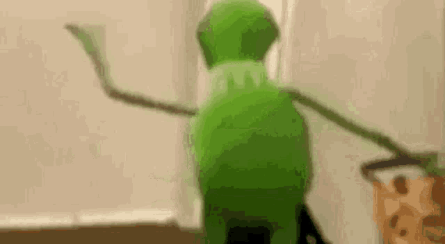 a green kermit the frog is standing in front of a window and holding a hockey stick .